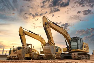 Construction Equipment on Site: Two heavy-duty excavators and a bulldozer stand ready for work on a construction site, silhouetted against a dramatic sunset sky.